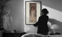 Mucha La Trappistine print in a slim black frame being hung on a sunlit wall in a monochrome room