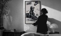 Nike x KAWS Air Force 1 print in a slim black frame being hung on a sunlit wall in a monochrome room