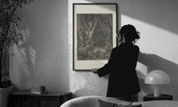 Dore Wood of the Harpies print in a slim black frame being hung on a sunlit wall in a monochrome room