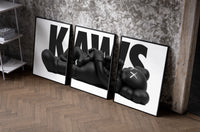 KAWS Monochrome Family Poster Set of 3 as black-framed prints leaning against a raw-plaster wall