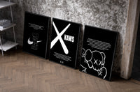 KAWS Monochrome Wall Art Set set of three black-framed prints leaning against a raw-plaster wall