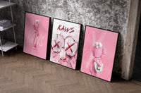 Bubblegum Pink KAWS XX Hypebeast Wall Art Set of 3 as black-framed prints leaning against a raw-plaster wall