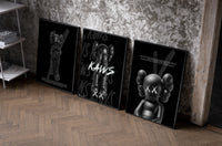 KAWS Companion Black Poster Set set of three black-framed prints leaning against a raw-plaster wall