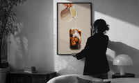 Iced Latte Cream Pour print in a slim black frame being hung on a sunlit wall in a monochrome room
