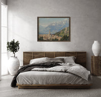 Alpine town with church tower print in a dark wood frame above a rattan bed on a plaster wall