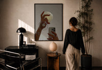 Jeweled Hands with Phone and Glass print in a black frame on a sunlit plaster wall, a woman looking on beside a mushroom lamp