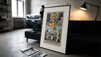 Angel blowing trumpet over rising figures print in a slim black frame leaning on a dark sofa in a concrete print studio