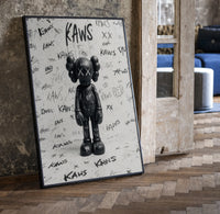 Black and white KAWS graffiti framed poster print in a black frame leaning beside a vintage door on parquet