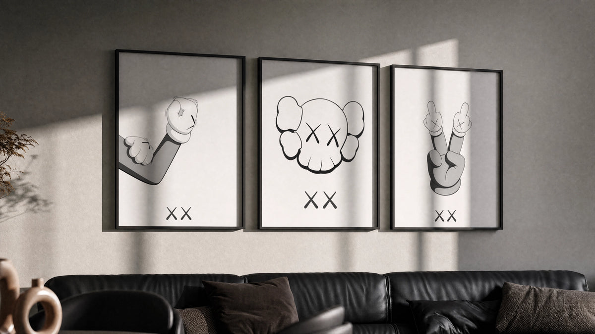 KAWS cartoon hands and skull face triptych in three black frames on a grey wall above a leather sofa