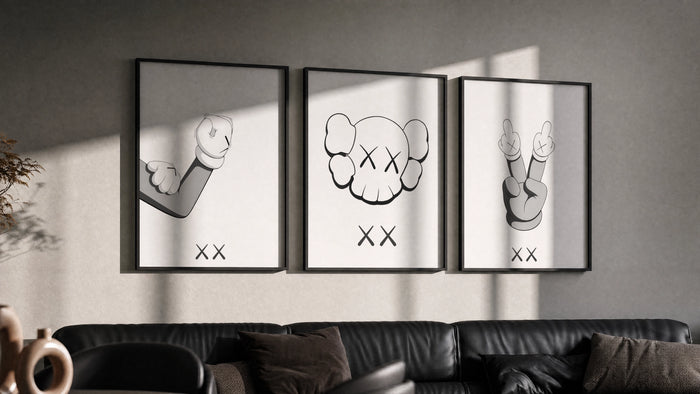 KAWS cartoon hands and skull face triptych in three black frames on a grey wall above a leather sofa