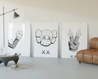 KAWS cartoon hands and skull face three-panel set displayed beside a brown leather sofa