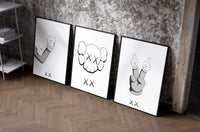 KAWS cartoon hands and skull face set of three black-framed prints leaning against a raw-plaster wall