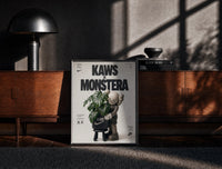 Black-framed KAWS Companion figure holding monstera plant print on a moody console with designer coffee-table books