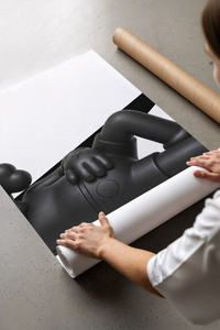 A panel of the Black stylized figure lying down with crossed eyes set being unrolled on a concrete floor