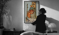 King of Wands print in a slim black frame being hung on a sunlit wall in a monochrome room