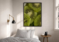 Kiwi Fruit Slices print in a black frame above a bed with striped linens, a brass wall lamp and a sunlit window