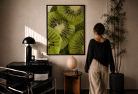 Kiwi Fruit Slices print in a black frame on a sunlit plaster wall, a woman looking on beside a mushroom lamp