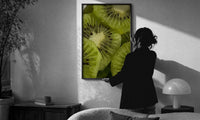 Kiwi Fruit Slices print in a slim black frame being hung on a sunlit wall in a monochrome room