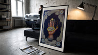 Upraised hand with alchemical symbols print in a slim black frame leaning on a dark sofa in a concrete print studio