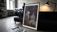 Old alchemist reading book with flask print in a slim black frame leaning on a dark sofa in a concrete print studio