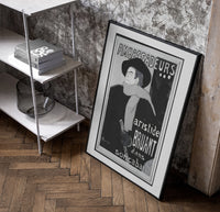Man in hat and scarf poster in a black frame leaning beside a shelf in a raw-plaster loft