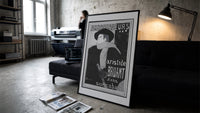 Man in hat and scarf print in a slim black frame leaning on a dark sofa in a concrete print studio