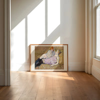 Two reclining women on sofa print in an oak frame on a sunlit wood floor beneath tall window light