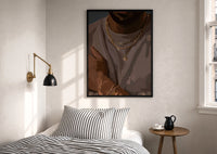 Layered Gold Chains on Taupe print in a black frame above a bed with striped linens, a brass wall lamp and a sunlit window