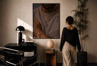 Layered Gold Chains on Taupe print in a black frame on a sunlit plaster wall, a woman looking on beside a mushroom lamp