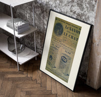 Vintage French soap advertisement with cat poster in a black frame leaning beside a shelf in a raw-plaster loft
