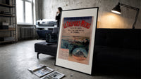 Coastal seaside town harbour view print in a slim black frame leaning on a dark sofa in a concrete print studio
