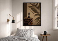 Leopard Print Chess King print in a black frame above a bed with striped linens, a brass wall lamp and a sunlit window