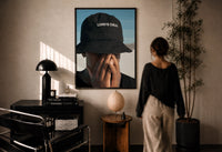 Lord's Child Bucket Hat print in a black frame on a sunlit plaster wall, a woman looking on beside a mushroom lamp