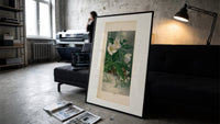 White hellebore flowers with sparrows print in a slim black frame leaning on a dark sofa in a concrete print studio