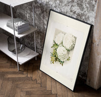 White chrysanthemum flowers with leaves poster in a black frame leaning beside a shelf in a raw-plaster loft