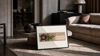 Purple pansy and pink blossom bouquet print in a slim black frame leaning on a travertine coffee table