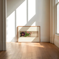 Purple pansy and pink blossom bouquet print in an oak frame on a sunlit wood floor beneath tall window light