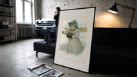 Blonde girl in plumed hat holding violets print in a slim black frame leaning on a dark sofa in a concrete print studio