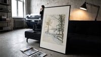 Hunter and dog in snowy landscape print in a slim black frame leaning on a dark sofa in a concrete print studio