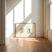 Two young girls holding flowers print in an oak frame on a sunlit wood floor beneath tall window light