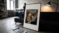 Two Victorian women looking at painting print in a slim black frame leaning on a dark sofa in a concrete print studio