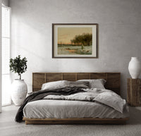 Winter landscape with frozen stream print in a dark wood frame above a rattan bed on a plaster wall