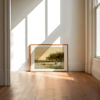 Winter landscape with frozen stream print in an oak frame on a sunlit wood floor beneath tall window light