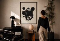 Melting 8 Ball print in a black frame on a sunlit plaster wall, a person looking on beside a mushroom lamp