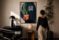 Mental Health Matters Potted Plant print in a black frame on a sunlit plaster wall, a woman looking on beside a mushroom lamp