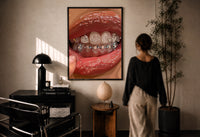 Mouth with Heart Tooth Gems print in a black frame on a sunlit plaster wall, a woman looking on beside a mushroom lamp