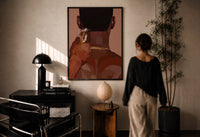 Neckline Trim with Clippers print in a black frame on a sunlit plaster wall, a woman looking on beside a mushroom lamp