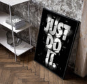 Just Do It typography design poster in a black frame leaning beside a shelf in a raw-plaster loft