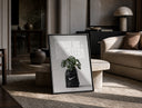 Monstera plant in Nike garbage bag print in a black frame leaning on a travertine table in a sunlit lounge