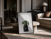 Monstera plant in Nike garbage bag print in a black frame leaning on a travertine table in a sunlit lounge